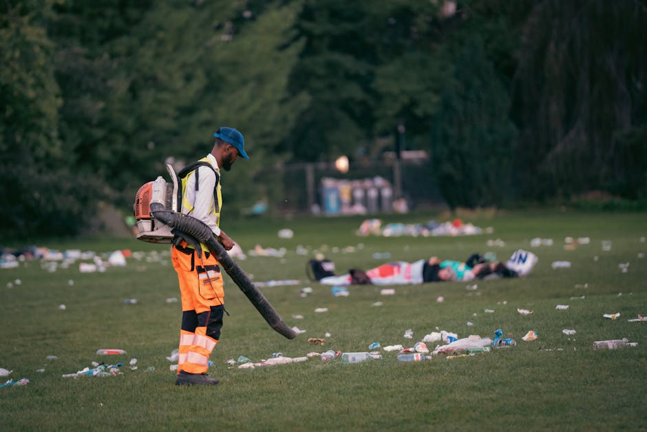 A waste management worker wearing a blue cap, reflective high-visibility jacket, and orange trousers with black knee patches is using a leaf blower to clear litter from a grassy field. The worker is p