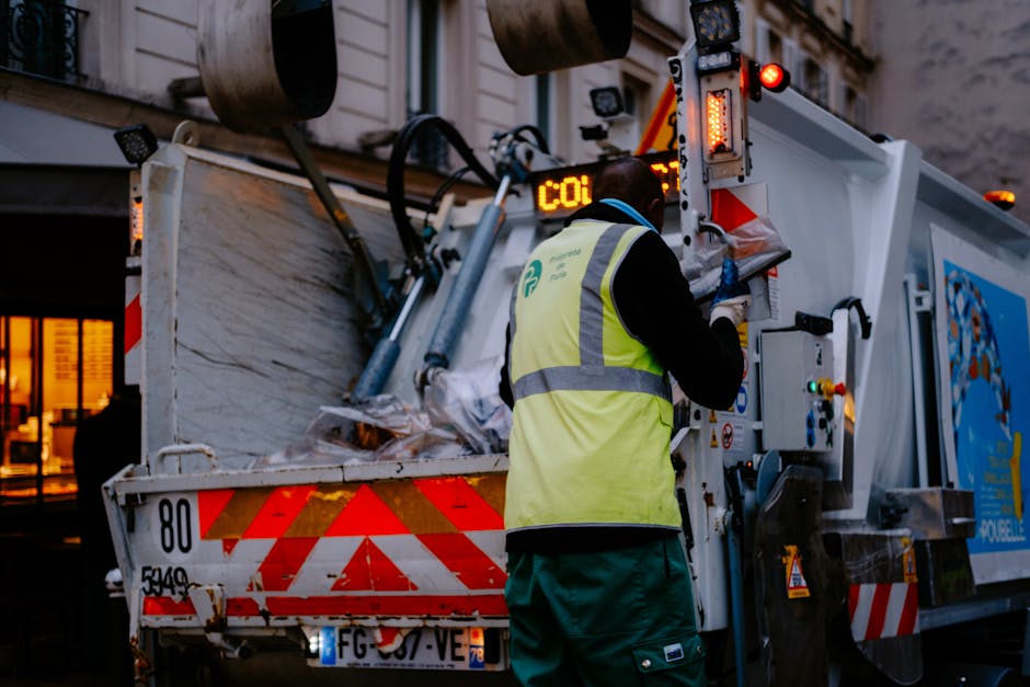 A waste management worker wearing a blue cap, reflective high-visibility jacket, and orange trousers with black knee patches is using a leaf blower to clear litter from a grassy field. The worker is p