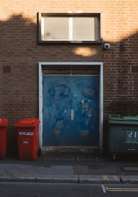 A professional waste collection worker in a high-visibility yellow and orange vest is standing next to a large red waste collection vehicle parked on the side of a street. The worker appears to be han